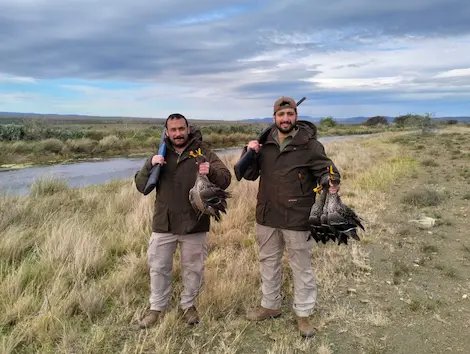 Two hunters standing next to a river posing with their ducks after a successfull geese hunting trip
