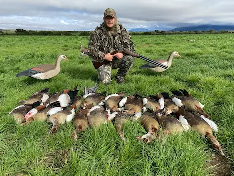 A hunter out in the pen green field posing with the ducks and geese he shot