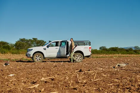 A Hunter standing in front of his truck in a field holding a shotgun