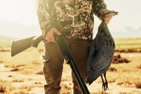 A hunter standing with a guineufowl in one hand, and a shotgun in the other