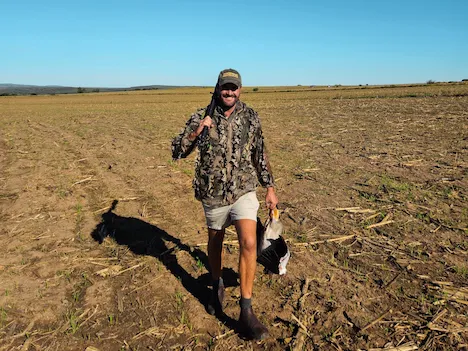 A smiling hunter walking over a field with a shotgun over his shoulder and a duck in his hand